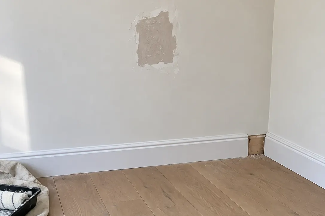 Room corner with a patched, unpainted plaster repair on the wall and a missing section of skirting board